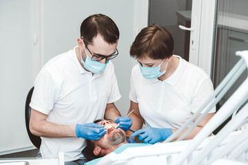 Obraz premium Dentist and his assistant treat a male patient in a dental clinic. Dentists in medical masks working with client's teeth. Copy, empty space for text