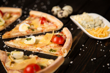 Slices of pizza with tomatoes, pesto sauce, bacon, quail eggs, mozzarella and Parmesan cheese on wooden background with sea salt and ingredients. Three types of cheese and quail eggs near pizza pieces