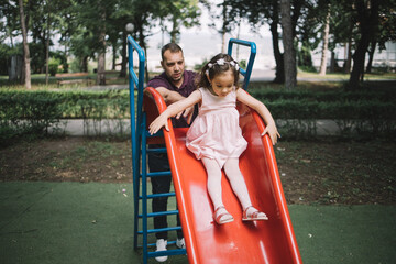 Little girl sliding down on slide in park. Handsome man helping little girl to slide on slider in...
