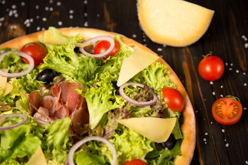 Part of Traditional Italian pizza with jamon Serrano, salad mix, cheese, black olives and cherry tomatoes on wooden background near half of tomato and piece of cheese. Tasty Mediterranean rustic meal.