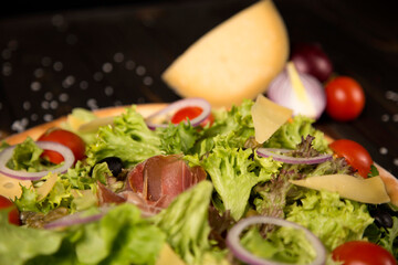 Close up of Traditional Italian pizza with jamon Serrano in focus, Parmesan cheese, black olives and cherry tomatoes on wooden background with ingredients. Tasty Mediterranean rustic food
