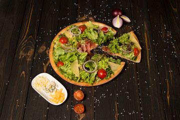 Side view of Italian pizza with jamon, Parmesan cheese, black olives, salad mix and cherry tomatoes on wooden background with sea salt and Ingredients near. Tasty pizza slice. Mediterranean food

