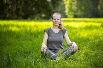 Naklejka na ściany i meble Yoga in the park. Woman in lotus position sits on the green grass in the park. The sun illuminates the grass in the background.