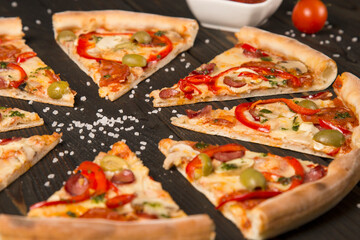 Slices of delicious pizza. Side view of pizza with bell pepper, olives, sausages, mozzarella cheese, pepperoni, salami, chili pepper, champignons on wooden background with sea salt in focus.
