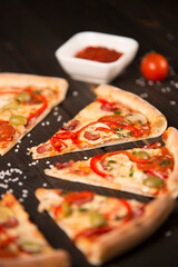 Half of sliced pizza with bell pepper, olives, sausages, mozzarella cheese, pepperoni, salami, chili pepper, champignons on wooden board with sea salt in focus and ingredients on background.
