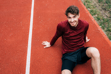 Obraz premium Handsome sportsman smiling at camera while sitting on running track outdoors
