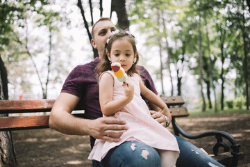 Cute little girl with headband eating ice cream in nature. Daughter looking at her ice cream while sitting in her father's lap in forest.