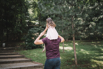 Naklejka premium Back view of father having daughter on his shoulders. Dad carrying his little girl on shoulders while walking in forest.