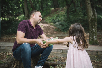 Fototapeta premium Dad and small girl holding present in park. Father sitting on stump in park giving present to his little daughter.