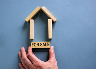 Male hand builds a model of a wooden house from wooden blocks. Words 'for sale'. Copy space. Business concept. Beautiful blue background.