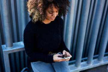 Afro american young pretty female with curly hair sitting outdoors holding modern smartphone booking hotel and transferring money in online banking service using 4G internet connection on weekends