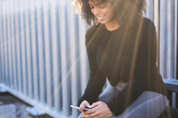 Smiling young Afro-American traveler making reportage about her trip writing about city in her blog typing text on modern smartphone connected to free wireless connection while sitting at street