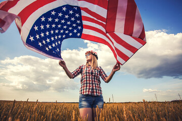 Beautiful young girl holding an American flag on the wind in a field of wheat. Summer landscape against the blue sky. Independence Day Fourth of July