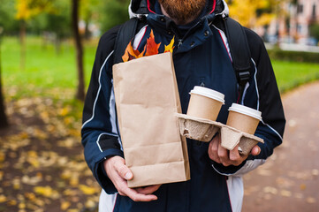 A young bearded caucasian man in a blue and black sports jacket with white stripes holds in his hands two paper cups and a paper bag with colorful maple leaves on a park background.
