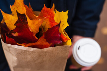 Crimson, orange and yellow maple leaves in a paper bag on the background of a hand with a paper cup with coffee.