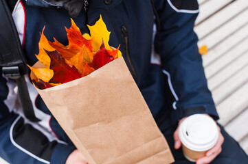 A paper bag with colorful maple leaves in the hands of a lonely-sitting caucasian man holding a paper cup in his left hand. A man is wearing a sports jacket.