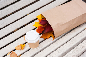 A paper cup with a white lid stands on a white old bench next to a paper bag, inside which are colored leaves of maple. Flat lay. Top view