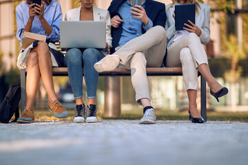 businesspeople sitting on bench outdoor with focus on legs, checking their cell phones, laptop,...