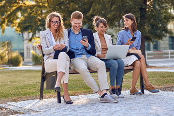 young caucasian people laughing together in park, sitting on bench, holding their cell phones,...