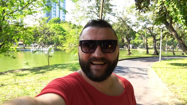 Portrait of happy man taking selfie, walking in city park, slow motion