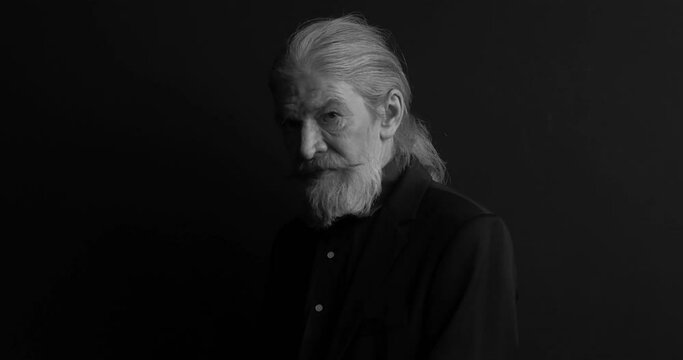 Close up. Old man with long hair and beard in dark business looking at camera and smile clothes standing sideways in semi-lit room on black. Black and white portrait. Waist portrait. Prores 422. 