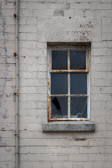 Rusted, decaying six pane metal window with cracked, broken glass recessed into dirty, white painted brick wall with concrete sill below and a vertical electrical cable clipped to wall