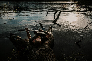 Europe, Germany, Berlin, Mueggelheim, Grosse Krampe, man relaxing on a tree in the lake with his guitar.