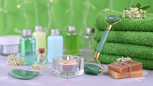 green facial jade roller among towel stack soap and candle against bottles with coloured liquids with white flowers closeup