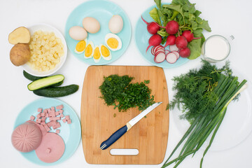 step by step recipe for Russian cold okroshka soup, ingredients for homemade okroshka cut with a knife-sausage, herbs, eggs, cucumbers, potatoes, radishes. top view on a light background