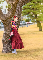 Obraz premium Japanese woman dressed in a hakama kimono and wearing a facial mask leaned against the trunk of a pine tree in the Tokyo Imperial Palace.