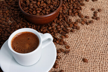 Espresso white cup, coffee beans, Arabica grains in bowl on jute burlap sackcloth. High angle, closeup, copy space. Coffee shop, morning, traditional roast concept