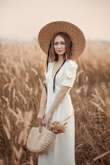 A beautiful Brunette girl in a light white dress and a straw boater hat is holding a straw bag.