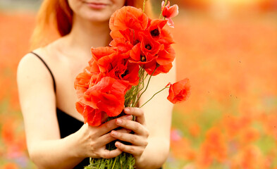 A young girl with red hair in a black sundress holds a bouquet with poppies in a poppy field outside the city. Summer and relaxation. A bouquet of red poppies. Summer concept. Flowers and floristry.