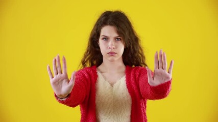 Serious attractive young woman with long wavy hair showing stop gesture with palm. Portrait of young successful confident girl isolated on yellow background. Concept of defense and lifestyle.