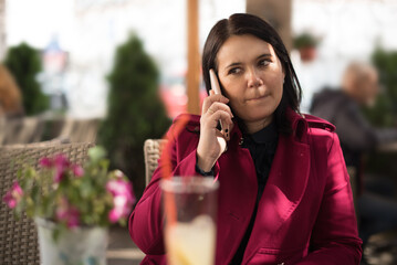 Image of a beautiful adult woman wearing a pink coat talking on a cellphone while sitting in 

street cafe restaurant outdoors