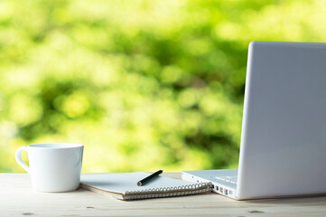 Workplace with white laptop, notebook and coffee on wooden desk ,which has a backdrop in a blurred natural green garden.