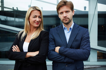 Two business colleagues standing against office building with brightly smile, experienced businessman standing with his female colleague smiling and looking to you, teamwork concept