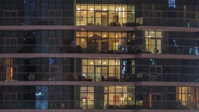 Rows of glowing windows with people in the interior of apartment building at night. Modern skyscraper with glass surface. Concept for business and modern life. Tilt down