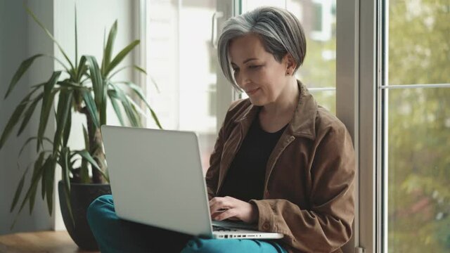 Charming gray haired mature woman working on laptop and look on her smartphone receive text message sitting leaning back against the window. Business concept. Prores 422.