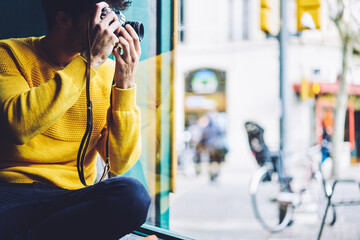 Talented creative young male photographer taking picture of city street to memorize weekend journey sitting inside of cozy coffee shop near window using unique vintage camera shooting black and white