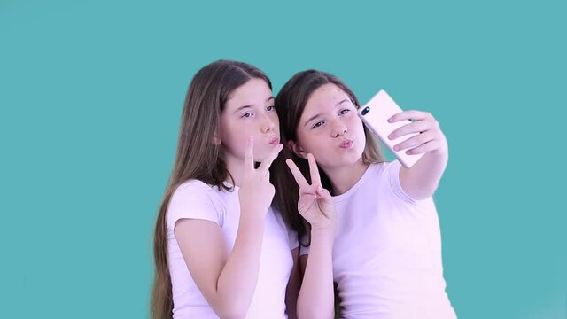 Cheerful caucasian sisters in white t-shirts are taking selfie with smartphone posing  making funny faces putting horns and checking their hair. Isolated on blue background. 