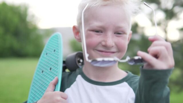 Close up a beautiful teenage boy with a skateboard in his hand. The boy takes off his sunglasses and smiles.