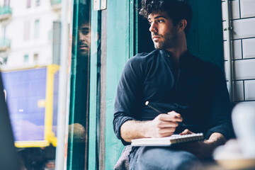 Toughtful hipster guy dressed in black shirt looking out of window and writing down text i notepad...