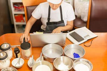 Female baker working on laptop with bakery ingredient on table.