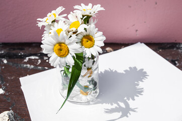 White blank sheet and a bouquet of daisies in a glass jar on a wooden background. Symbol of purity and freshness, skin cosmetics
