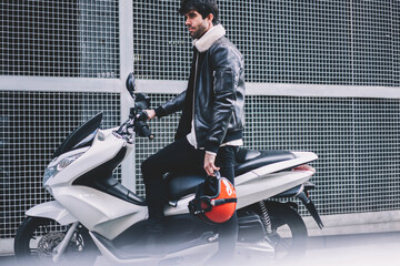 Confident bearded biker dressed in leather jacket standing near white motorcycle with powerful...