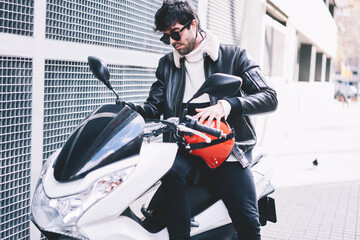 Experienced driver in stylish black sunglasses parking motorcycle on street.Cool hipster guy in spectacles sitting on white powerful motorbike with red helmet in urban setting