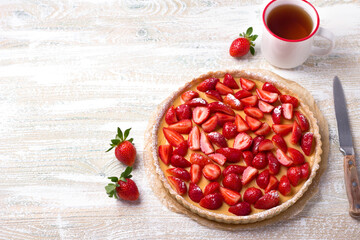 Summer tart with custard of baked milk, juicy fresh strawberries and a cup of tea on a light wooden background, top view, space, horizontal	