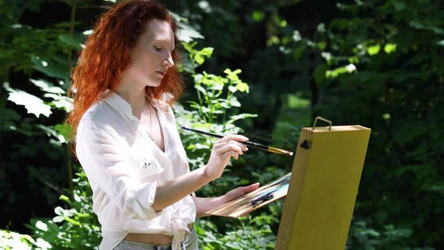 Middle aged redhead woman draws a picture in the open air in the forest
