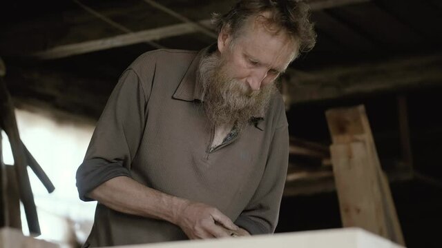 Male carpenter with a gray beard, works as a chisel. An elderly artisan will carry out his work intently in a workshop. Handwork of the master.